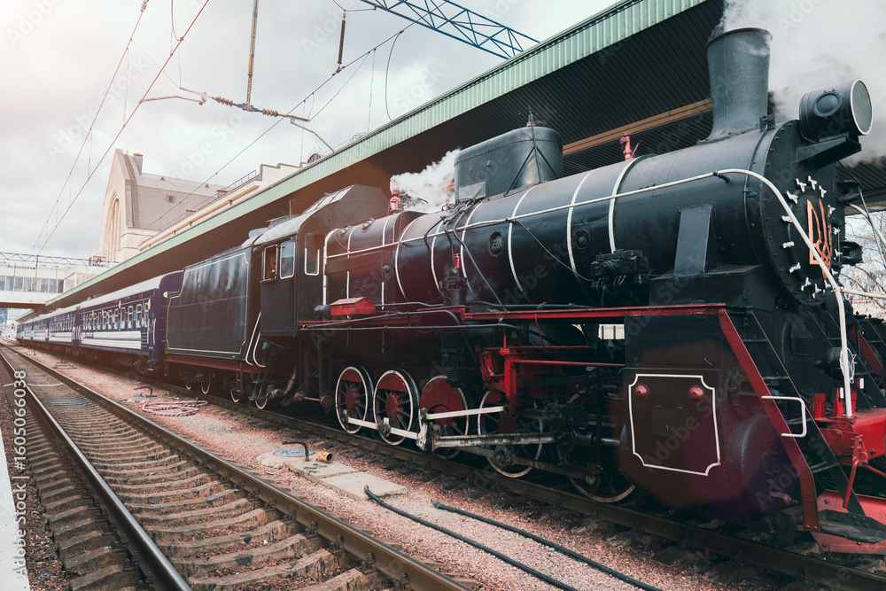 Naklejka premium A classic black steam train with red accents emits smoke while stopped at a railway station platform, evoking vintage travel nostalgia