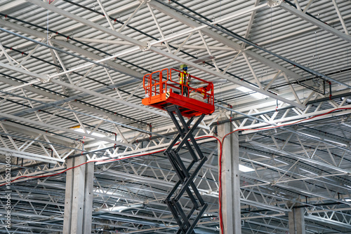 Worker on scissor lift platform inspecting or installing electrical systems on industrial warehouse ceiling with metal beams and lighting fixtures