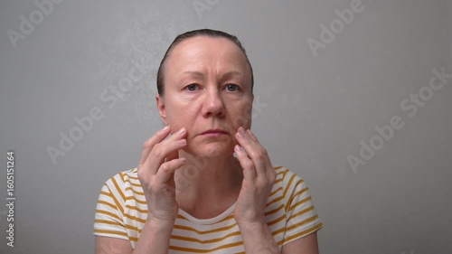 50-55-year-old woman looks with chagrin at her face with wrinkles, puffiness, and flabby skin. Age-related changes and fatigue. A woman is planning anti-aging procedures or plastic surgery