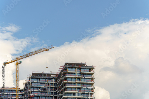 Fotografi Construction of a multi-unit residential building is underway with the help of a tower crane against a backdrop of blue sky and clouds