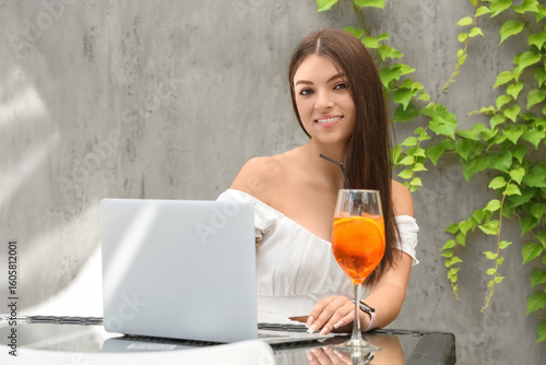 Beautiful young Asian woman with glass of tasty aperol spritz and laptop sitt...