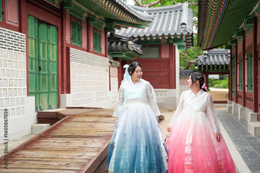 Fototapeta premium Seoul, June 2025: Two Korean women in their twenties stroll through Gyeongbokgung Palace's halls wearing pink and sky-blue hanbok. Bathed in summer light, they share laughter amid historic beauty.