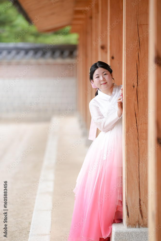 Fototapeta premium A Korean woman in her twenties wearing a charming pink hanbok peeks from the shadow of a wooden pillar at Gyeongbokgung Palace, her smiling face partially hidden by the column, in Seoul in June 2025.