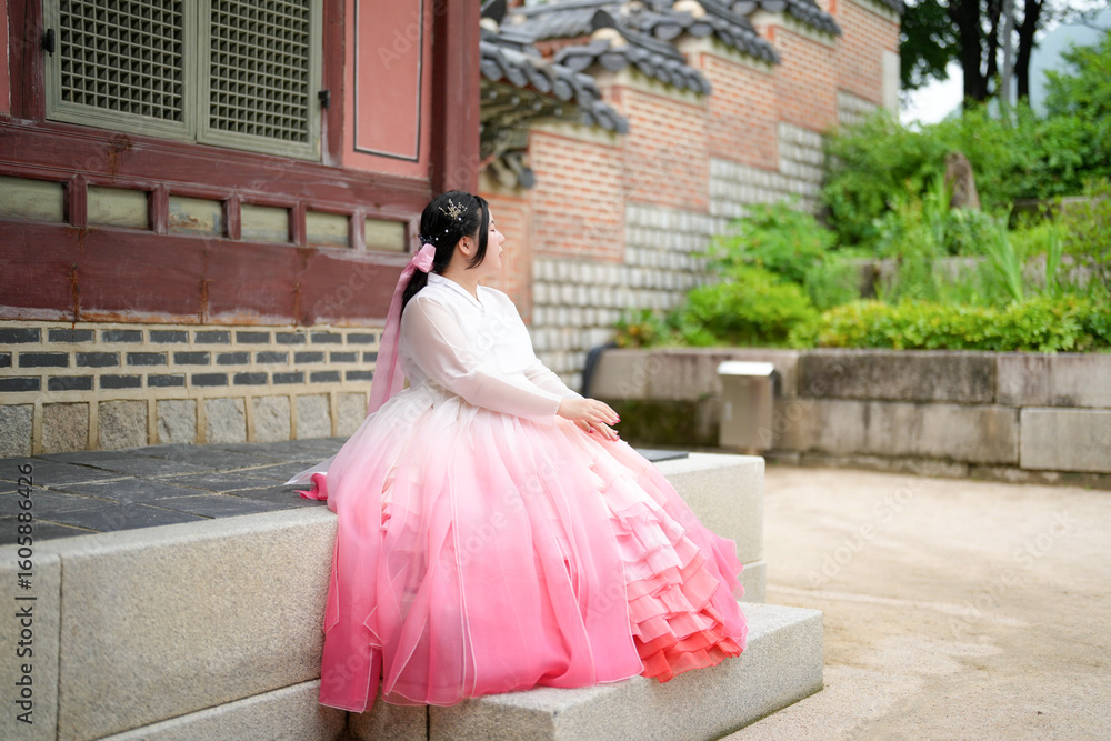 Fototapeta premium A Korean woman in her twenties wearing a charming pink hanbok sits in front of the traditional facade of Gyeongbokgung Palace, her calm demeanor reflecting timeless beauty, in Seoul in June 2025.