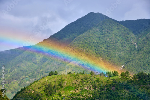 Vibrant rainbow arcs gracefully over lush green mountains, creating a stunning natural spectacle amidst serene landscape over Ulba Falls in Banos de Agua Santa, Ecuador, evoking sense of wonder