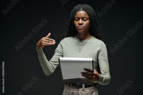 Young adult Black woman rehearsing lines during acting audition, holding script in one hand and gesturing with other hand, standing against plain dark background