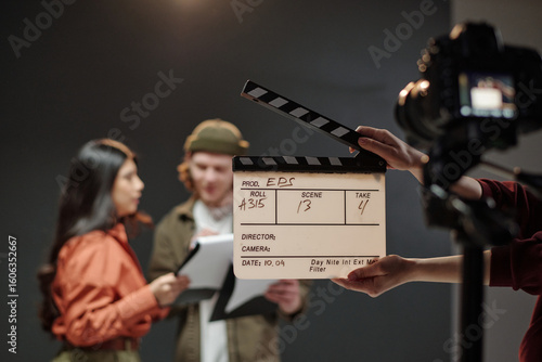 Young adult Caucasian man and young adult Hispanic woman standing together reading scripts during acting audition while camera and clapperboard visible in foreground capturing scene