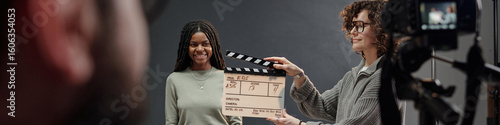 Black girl standing in front of camera smiling during acting audition while Caucasian woman holding clapperboard, professional film equipment visible in studio setting
