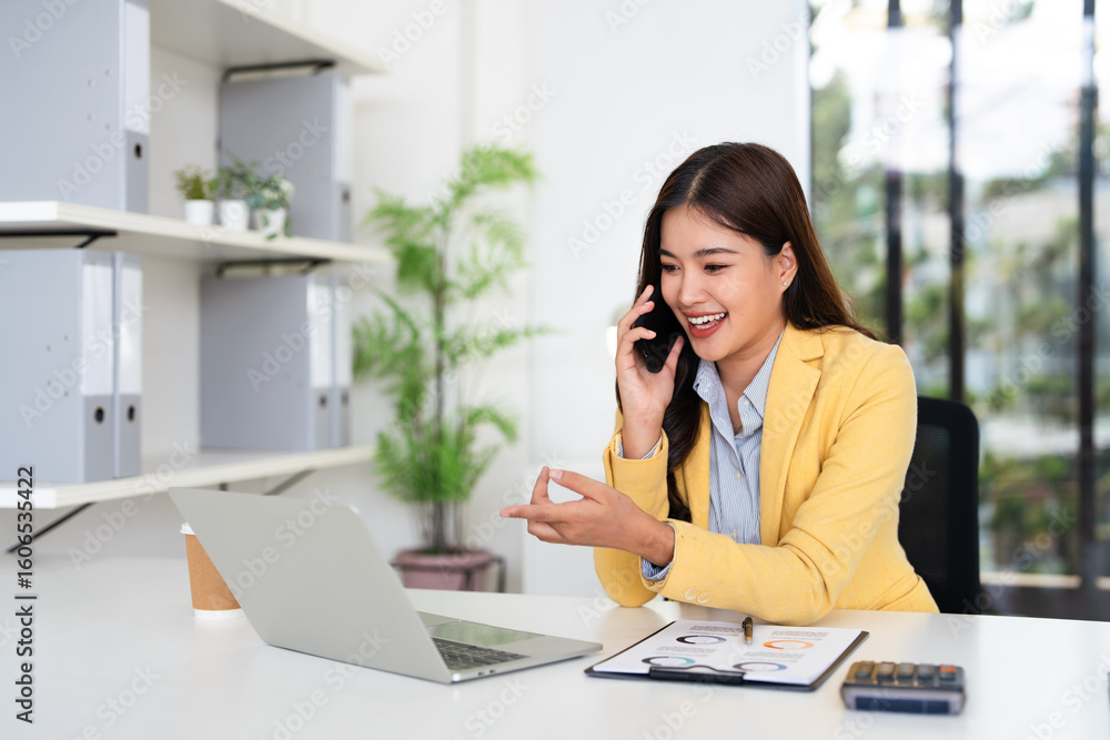 Fototapeta premium Happy joyful confident asian business lady, broker or officer, sitting at table in a modern office, working with laptop, talking to client or employees on smartphone.