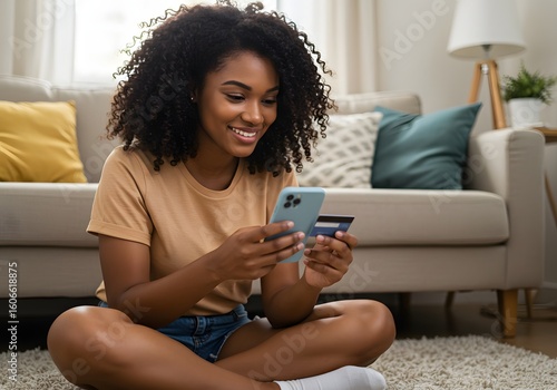 Young adult African American female consumer holding credit card and smartphone sitting on floor at home doing online banking transaction. E commerce virtual shopping, secure mobile banking concept.