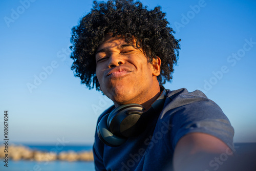 Playful young man pouting for a selfie at the beach with headphones around neck
