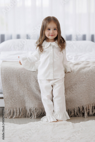 Child in white pajamas standing on bed beside cozy blanket in bright room