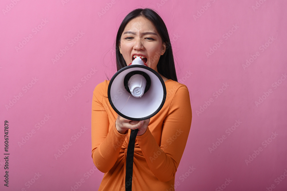 Fototapeta premium Asian woman with long black hair wearing an orange dress holding a megaphone directly facing the camera, standing against pink background, symbolizing voice, announcement, or call to action