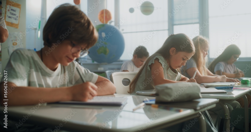 Fototapeta premium Elementary School Students Writing Exam or Doing Tasks in Notebooks. Female Teacher Walking Between Desks, Controlling Young Boys and Girls During School Test Process in Modern Classroom. Static Shot.