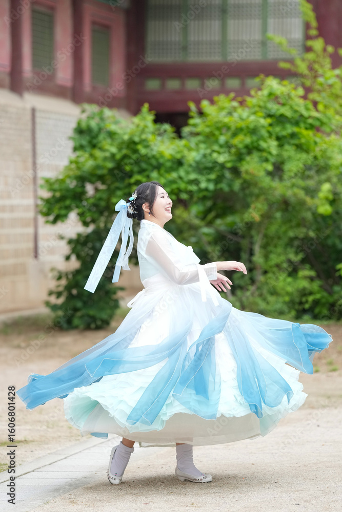 Fototapeta premium A Korean woman in her twenties wearing a sky-blue hanbok twirls gracefully in front of the traditional facade of Gyeongbokgung Palace, her skirt fanning out in elegant ripples, in Seoul in June 2025.