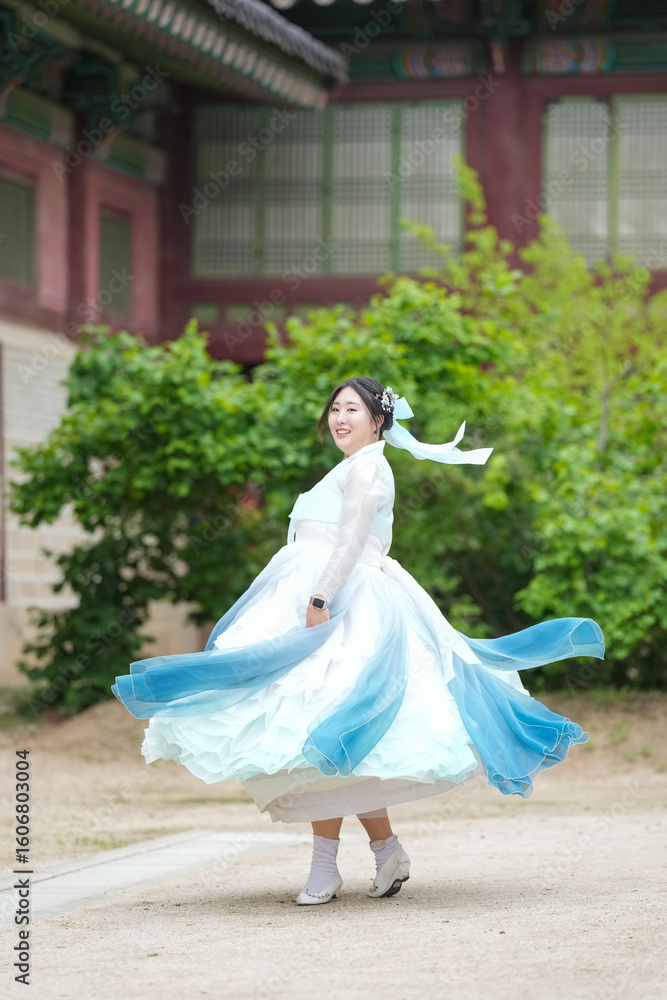 Fototapeta premium A Korean woman in her twenties wearing a sky-blue hanbok twirls gracefully in front of the traditional facade of Gyeongbokgung Palace, her skirt fanning out in elegant ripples, in Seoul in June 2025.