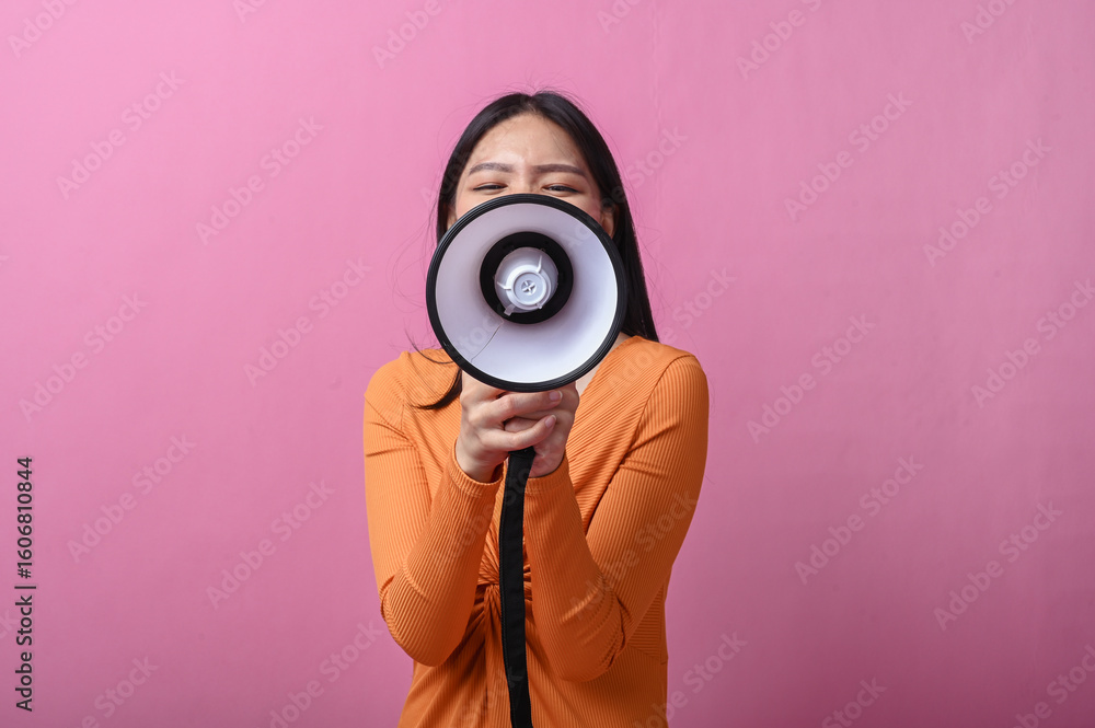 Fototapeta premium Asian woman with long black hair wearing an orange dress holding a megaphone directly facing the camera, standing against pink background, symbolizing voice, announcement, or call to action
