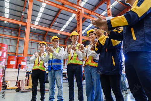 Group of industrial workers in safety vests and helmets clapping hands in celebration at warehouse. Positive atmosphere shows teamwork and employee appreciation in modern factory.
