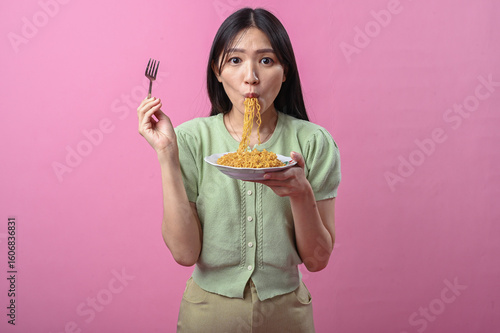 Asian woman in green top eating instant noodles with a fork from a white plate, enjoying her food against a vibrant pink background