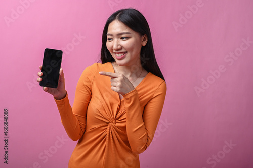 Asian woman with long black hair in orange dress smiling and pointing at smartphone screen with index finger, standing indoors against pink background, promoting or showing mobile content