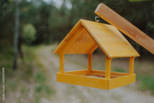 Papier peint Close-up of a bright yellow wooden bird feeder hanging outdoors with a forest path in the blurred background