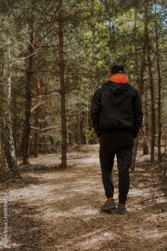 Wallpaper Mural Man in a black jacket and orange hoodie walks alone through a peaceful forest trail, surrounded by tall trees and soft natural light, viewed from behind. Torontodigital.ca