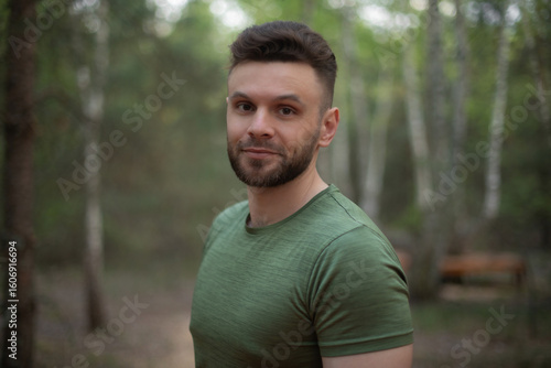 Portrait of a smiling young man in a green t-shirt standing in a birch forest. Natural light and soft background create a warm and friendly outdoor atmosphere.