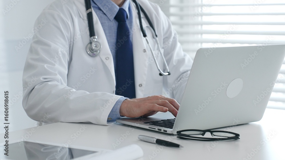 Male doctor in lab coat and stethoscope sitting at desk typing on laptop, holding glasses and pen nearby in medical office. Medicine and healthcare concept
