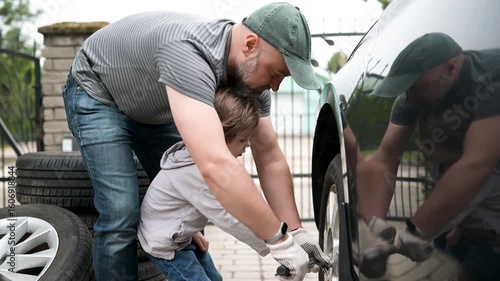 Cute toddler boy helping his father to change car wheels in their backyard. Father teaching his little son to use tools. Active parent of a small child. Slow motion summer day footage.