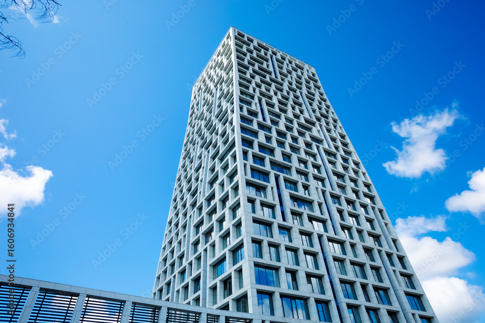 Modern building with geometric facade against blue sky, clouds. Sunny day emphasizes height, style, windows, lines, structure. Tall skyscraper rushes into clear sky Modern building with geometric facade against blue sky, clouds. Sunny day emphasizes height, style, windows, lines, structure. Tall skyscraper rushes into clear sky