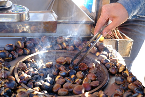 Chestnuts, a traditional Turkish street food. A close-up of a hand holding a chestnut with tongs. Smoke rises as the chestnuts roast over a charcoal fire. Winter.
