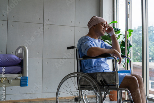 Asian man cancer patient sitting alone in wheelchair looking outside window in hospital ward with feeling of stress and lonely, health and medical care concept.