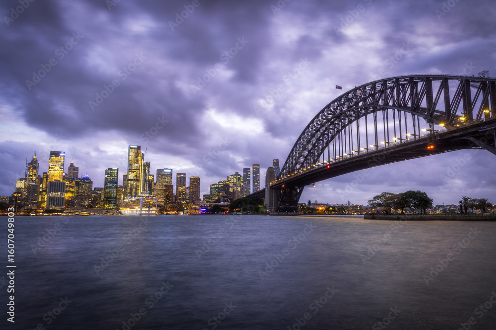 Sydney, Australia - 21 May 2025: View of the iconic Harbour Bridge arching gracefully against the city skyline, its lights glimmering on the tranquil water under a brooding sky. Sydney, Australia - 21 May 2025: View of the iconic Harbour Bridge arching gracefully against the city skyline, its lights glimmering on the tranquil water under a brooding sky.