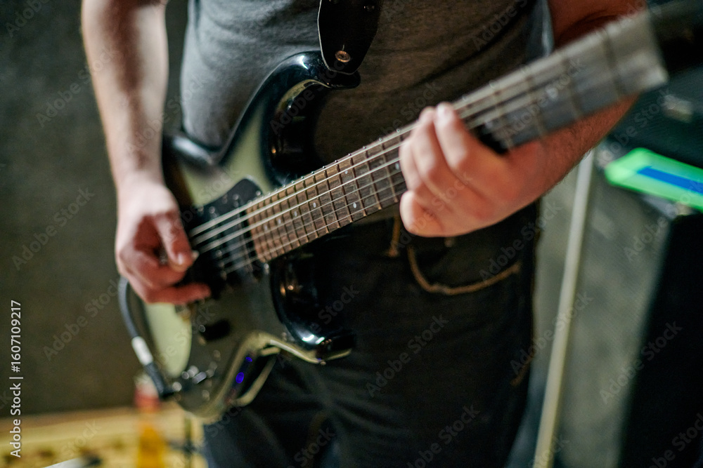 Obraz premium Musician playing an electric bass guitar at music rehearsal, practicing guitar technique in dimly lit setting, close up view guitarist hands