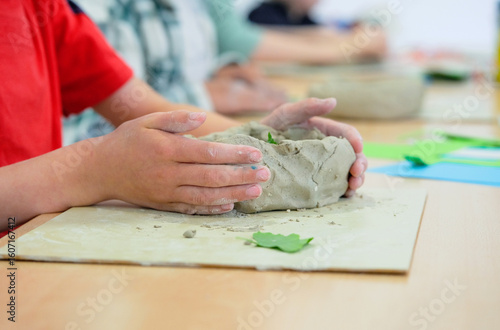 Detailblick auf die Hände eines Jungen beim kreativen Basteln mit Ton, Lehm oder Knetmasse an einem Tisch mit vielen bunten Materialien, im verschwommen Hintergrund  sind weitere Kinder