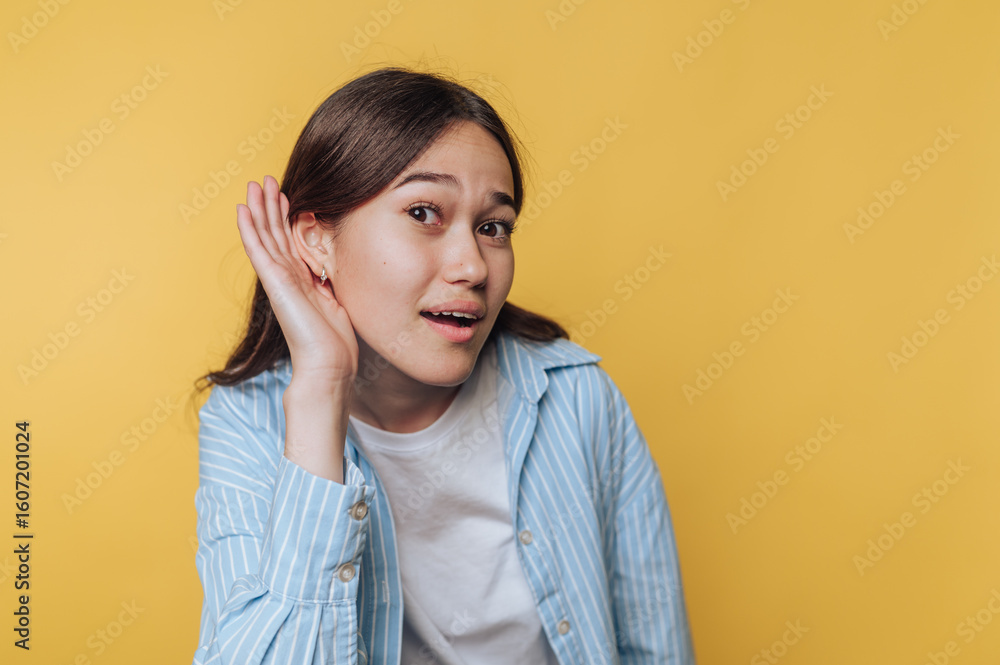 Young woman in a striped shirt holding her hand to her ear, listening intently against a yellow background. Young woman in a striped shirt holding her hand to her ear, listening intently against a yellow background.