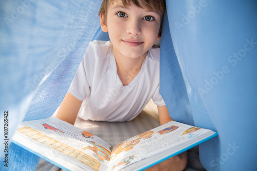 Smiling boy reading a book under a fabric tent