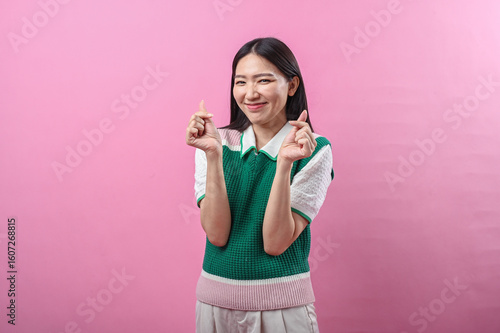 Asian woman smiling with both hands making Korean finger heart gesture, wearing a green knit vest, standing against a solid pink background