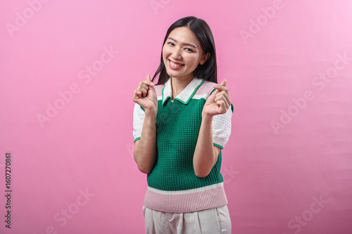 Asian woman smiling with both hands making Korean finger heart gesture, wearing a green knit vest, standing against a solid pink background