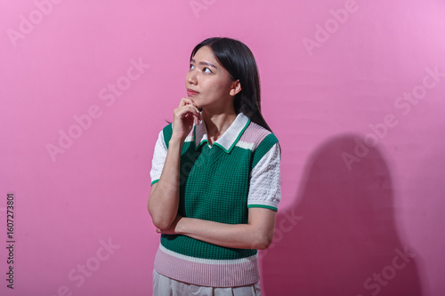 Asian woman in a green and pink sweater vest with a thoughtful expression, eyes closed and hand on chin, standing in front of a plain pink background