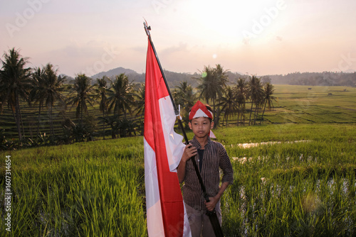 A boy in traditional Indonesian style carrying a red and white flag on a stick in the middle of a rice field with the sun setting in the open air