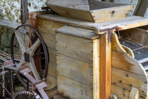 Фотография Old wooden threshing machine with wheels and mechanical drive, displayed against a rural building backdrop