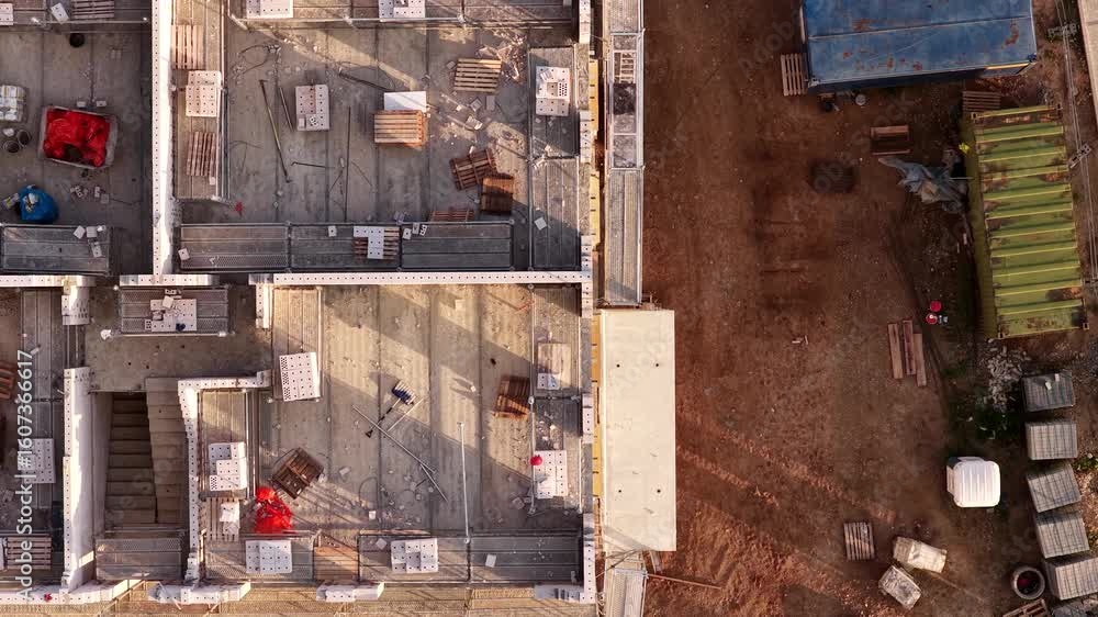 Overhead drone image of a multi-room structure under construction, showing scaffolding, bricks, pallets, stair access, and surrounding construction site equipment.