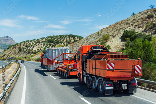 Heavy haul trucks transporting a massive metal crate on a steep incline, with one truck in front and another behind the load. Rear view.