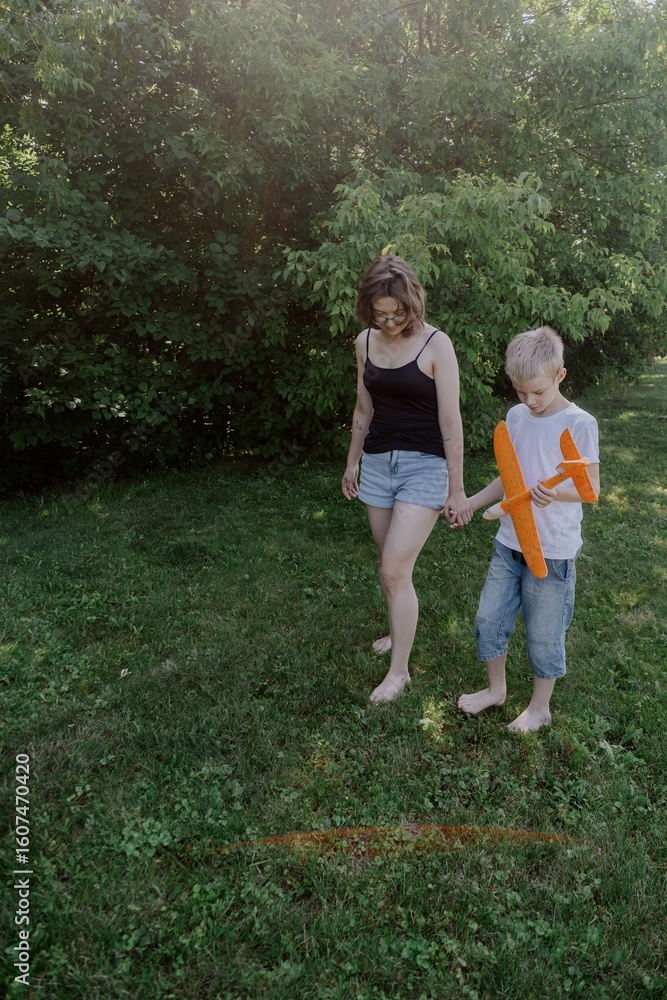 Fototapeta premium A mother and her son with hearing aids launch a paper airplane in a park, a heartwarming moment of inclusive play and bonding in nature. Perfect for disability awareness and family lifestyle content