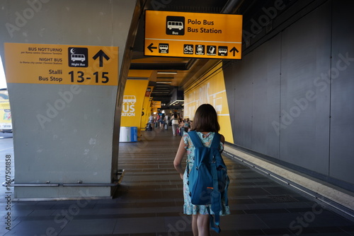 At Fiumicino Airport  a young girl with a blue backpack walks toward the brightly signed Polo Bus Station area, heading to bus stops for city and night routes - Blurred people in the background.