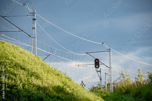 Modern railway traction system with red signal light, set against vibrant green grass and a dramatic cloudy sky. A clean blend of technology and nature captured in one frame