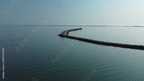 Aerial drone footage of a calm sea surrounding a breakwater with a coastal structure. Peaceful seascape with gentle water, rocks, and a maritime setting under natural light.