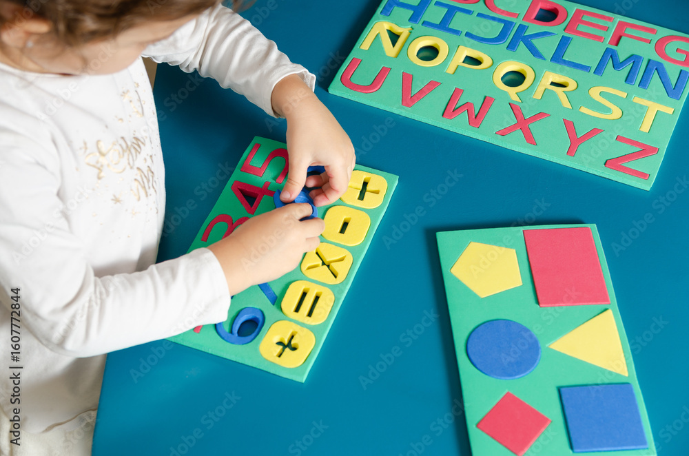 Obraz premium A little girl playing with colorful foam letters, numbers and geometric shapes on a blue table.
