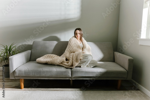 Cuadro en lienzo Young woman wrapped in chunky knit blanket on sofa